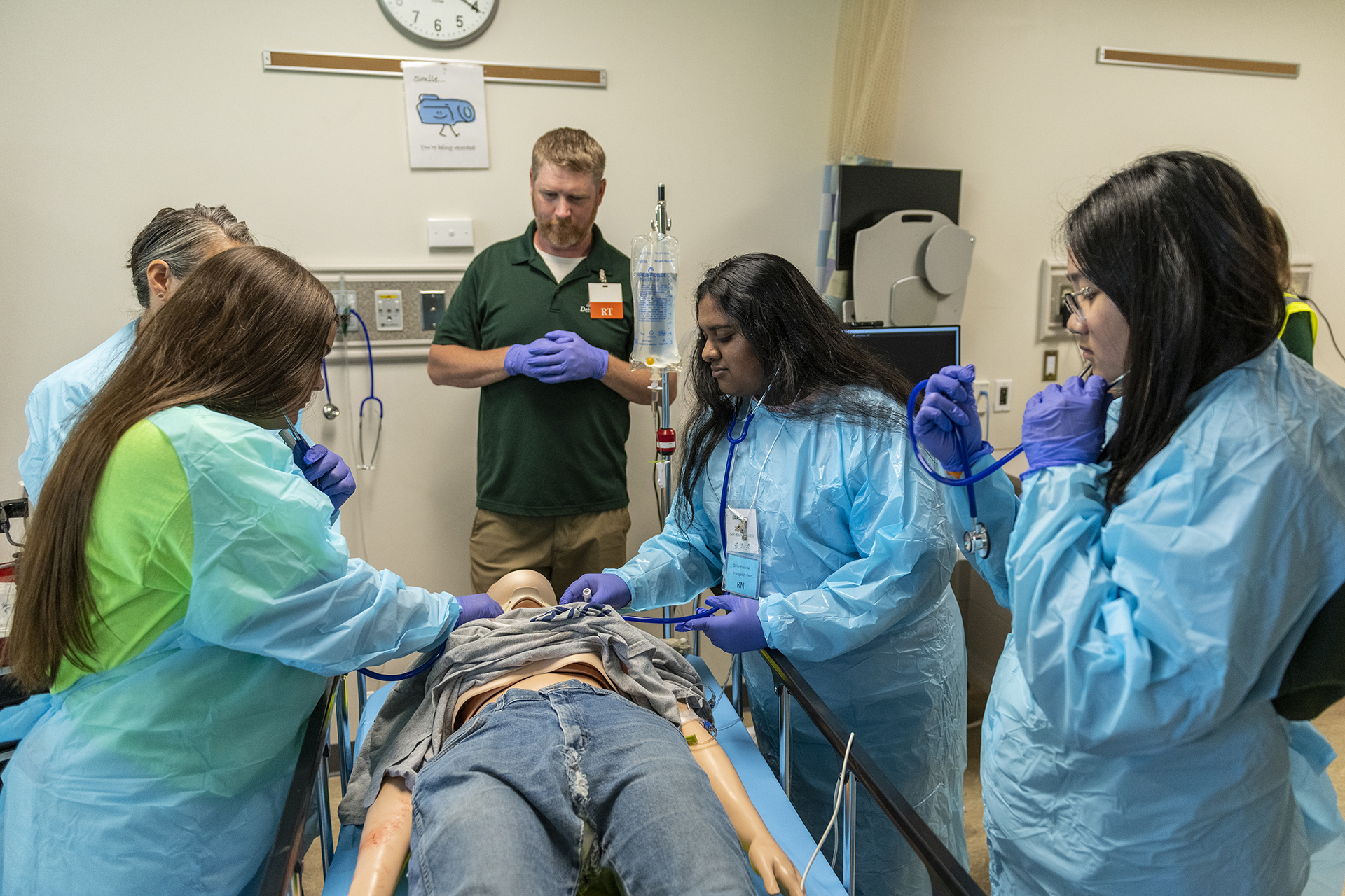 Students in blue surgical gowns stand around a stretcher with a mannequin