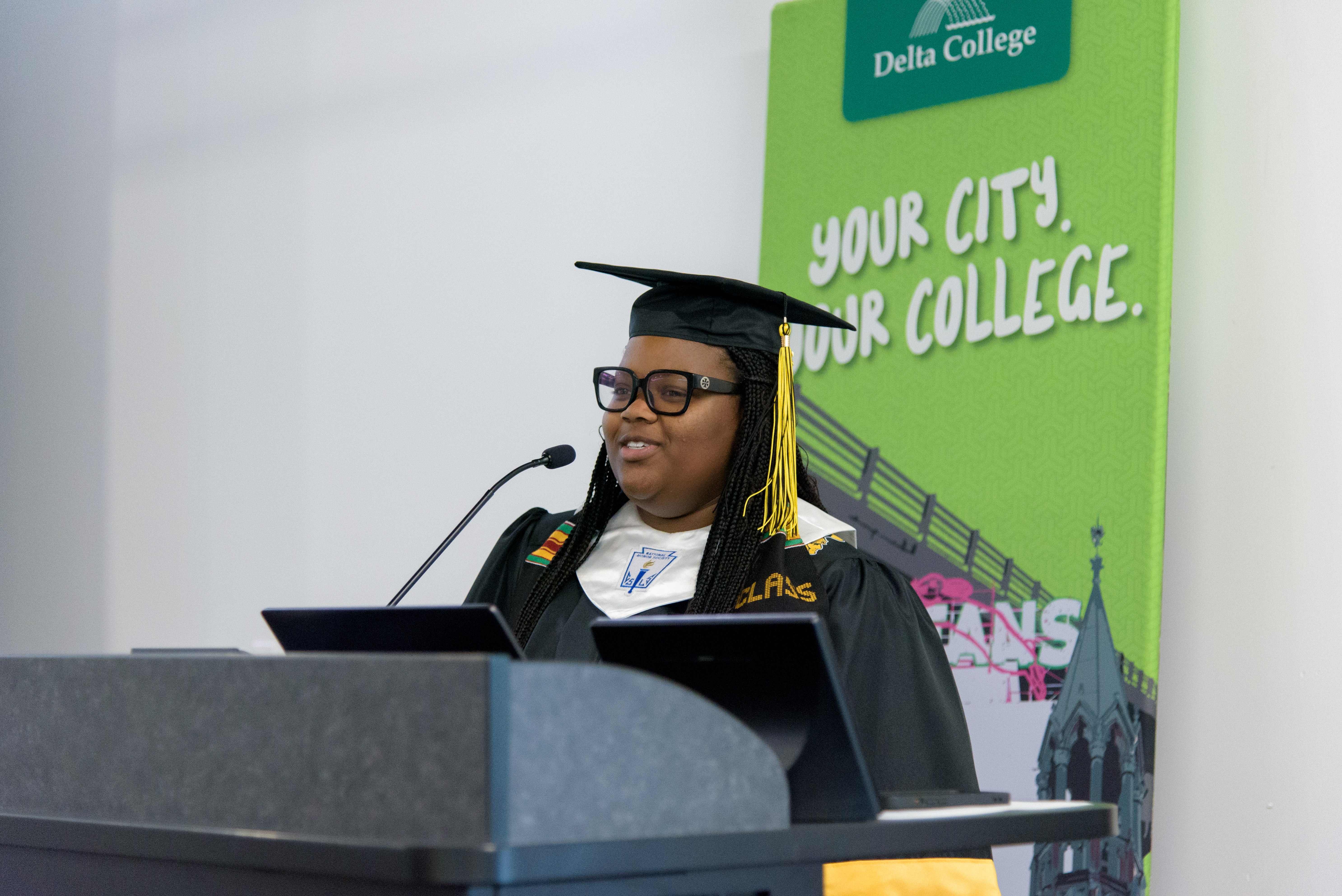 Tamariay Skinner wearing black cap and gown speaks in front of Delta College banner