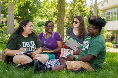 Four Delta College students sit in green grass on campus laughing together with notebooks in their hands and laps