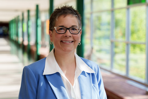 Shelly Raube female wearing dark-rimmed glasses, cropped hair, smiling wearing light blue blazer for a photo